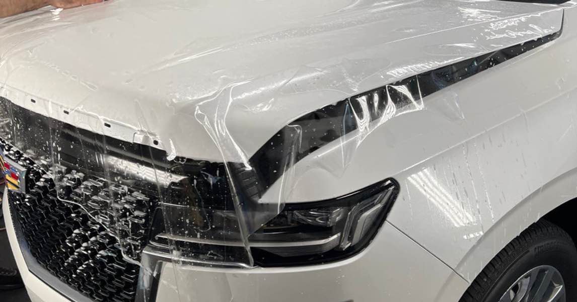 Technician applying clear paint protection film to a white Cadillac Escalade hood.